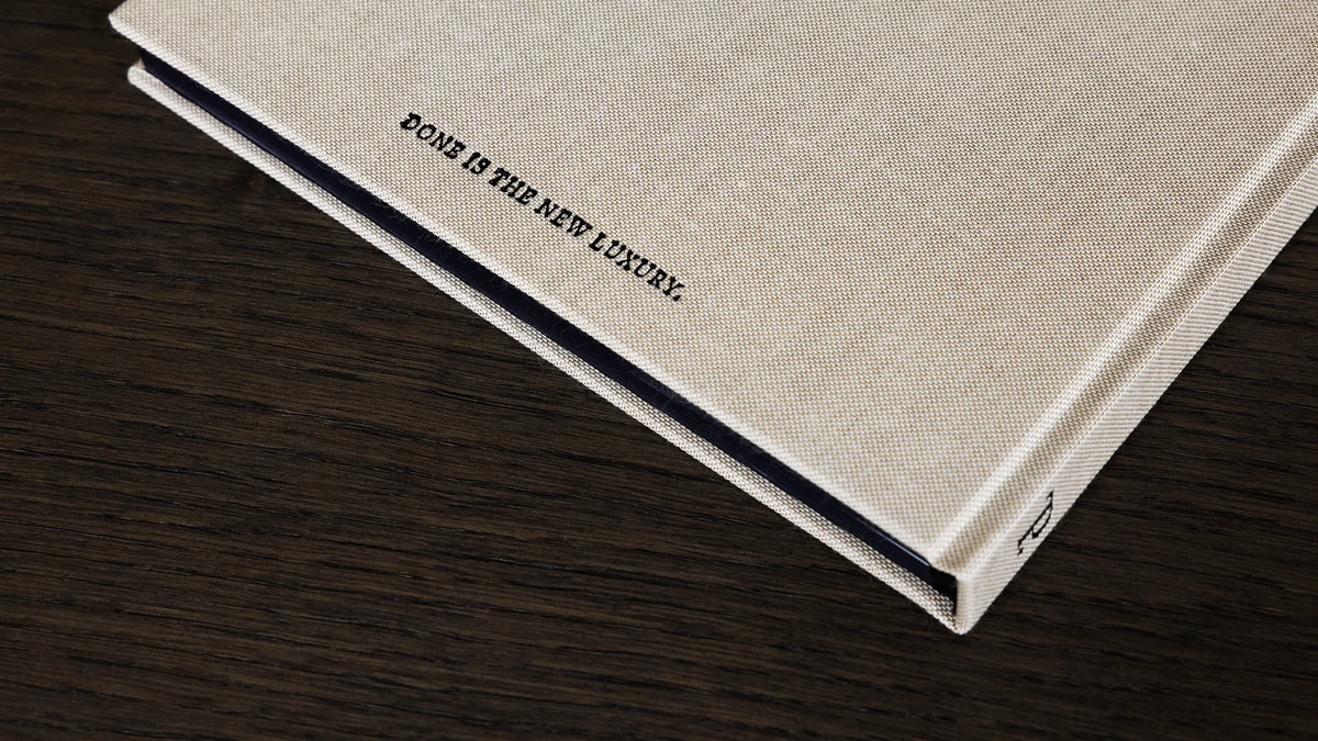 A close-up of a hardcover book - The PLNR - with a textured cream linen cover and matte black edging, resting on a dark wood surface. The back cover features the text 'DONE IS THE NEW LUXURY.